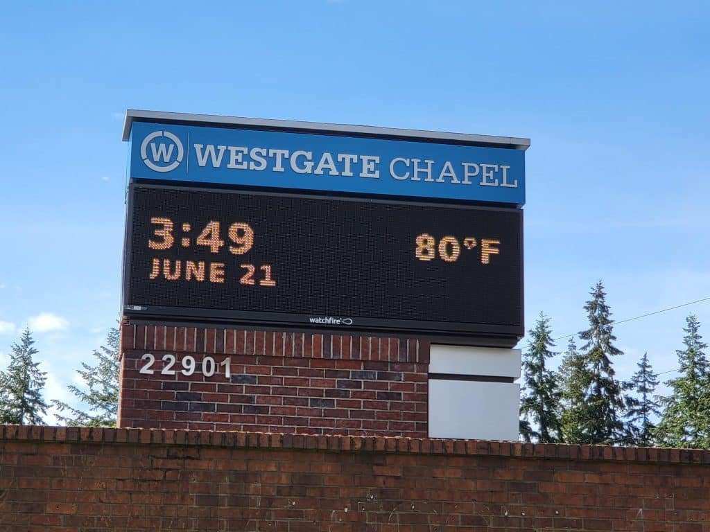 Electronic sign for Westgate Chapel displays the time 3:49, date June 21, and temperature 80°F, against a background of trees and a blue sky. Electronic sign for Westgate Chapel displays the time 3:49, date June 21, and temperature 80°F, against a background of trees and a blue sky.