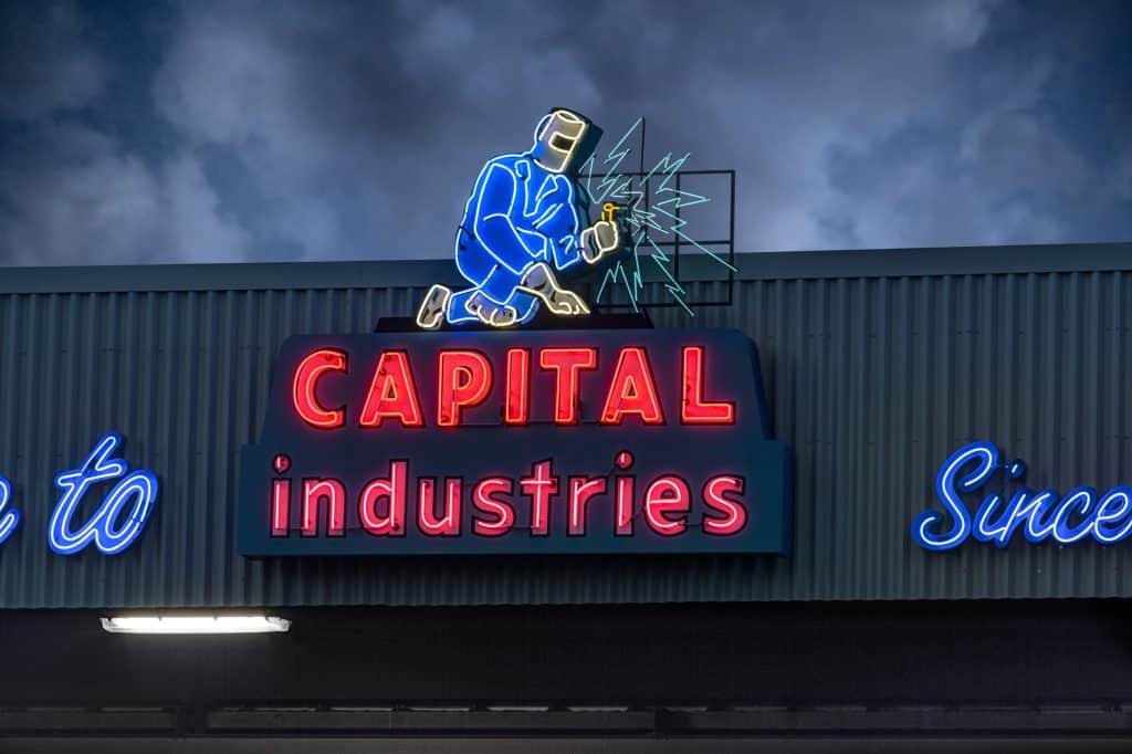 Neon sign depicting a welder above the words "CAPITAL industries" in red and blue on an industrial building. Neon sign depicting a welder above the words "CAPITAL industries" in red and blue on an industrial building.