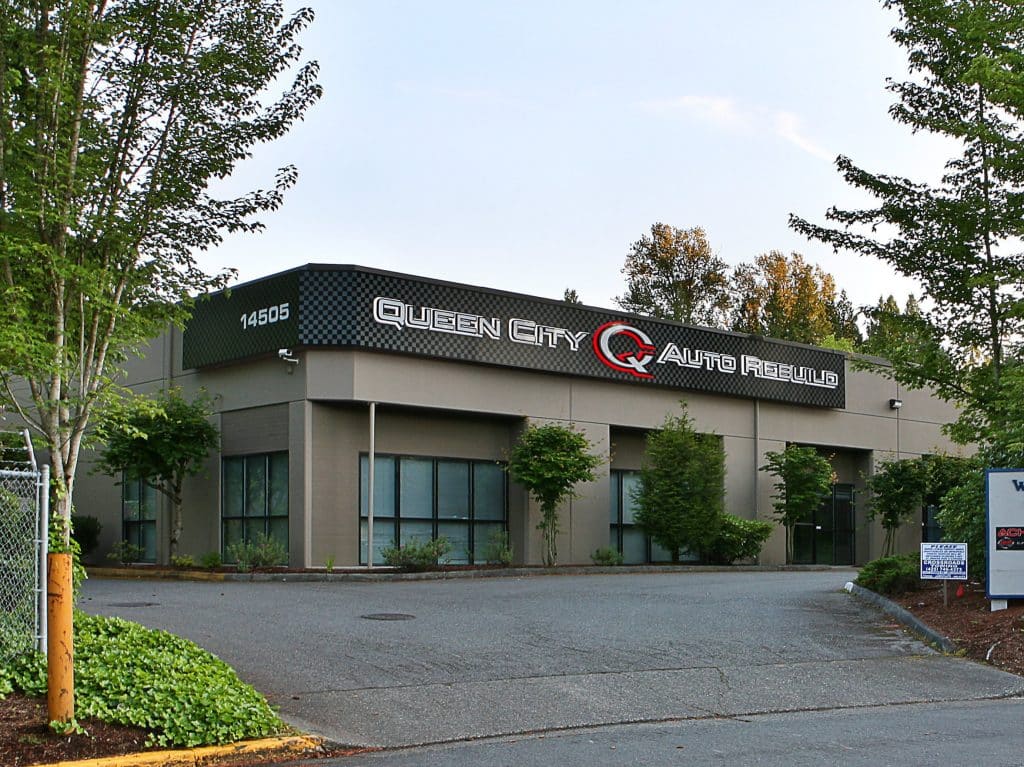 A large gray building labeled "Queen City Auto Rebuild" is surrounded by trees, with a parking lot in front.