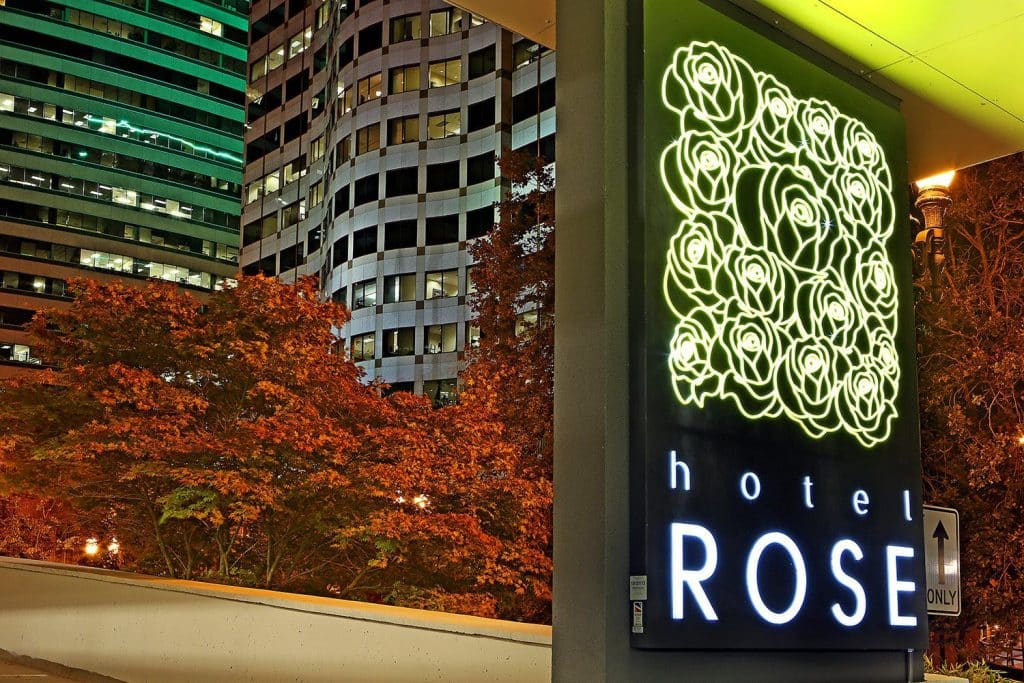 Neon sign for Hotel Rose with rose graphics, set against a backdrop of urban buildings and autumn trees at dusk. Neon sign for Hotel Rose with rose graphics, set against a backdrop of urban buildings and autumn trees at dusk.