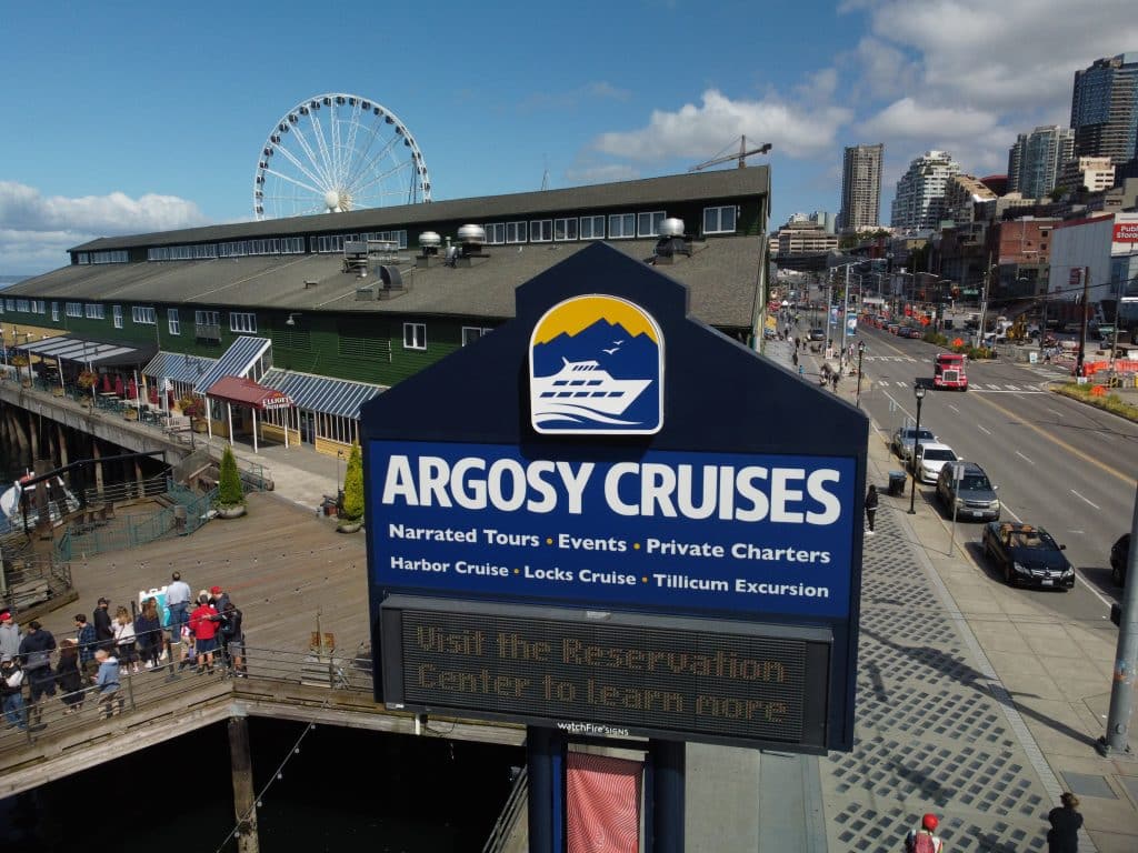 DCIM100MEDIADJI_0484.JPG Sign for Argosy Cruises in a waterfront area, with a Ferris wheel, building, and busy street in the background.