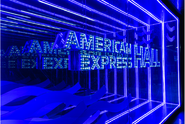 Blue neon sign reading "AMERICAN EXPRESS HALL" with reflections in a mirrored space.