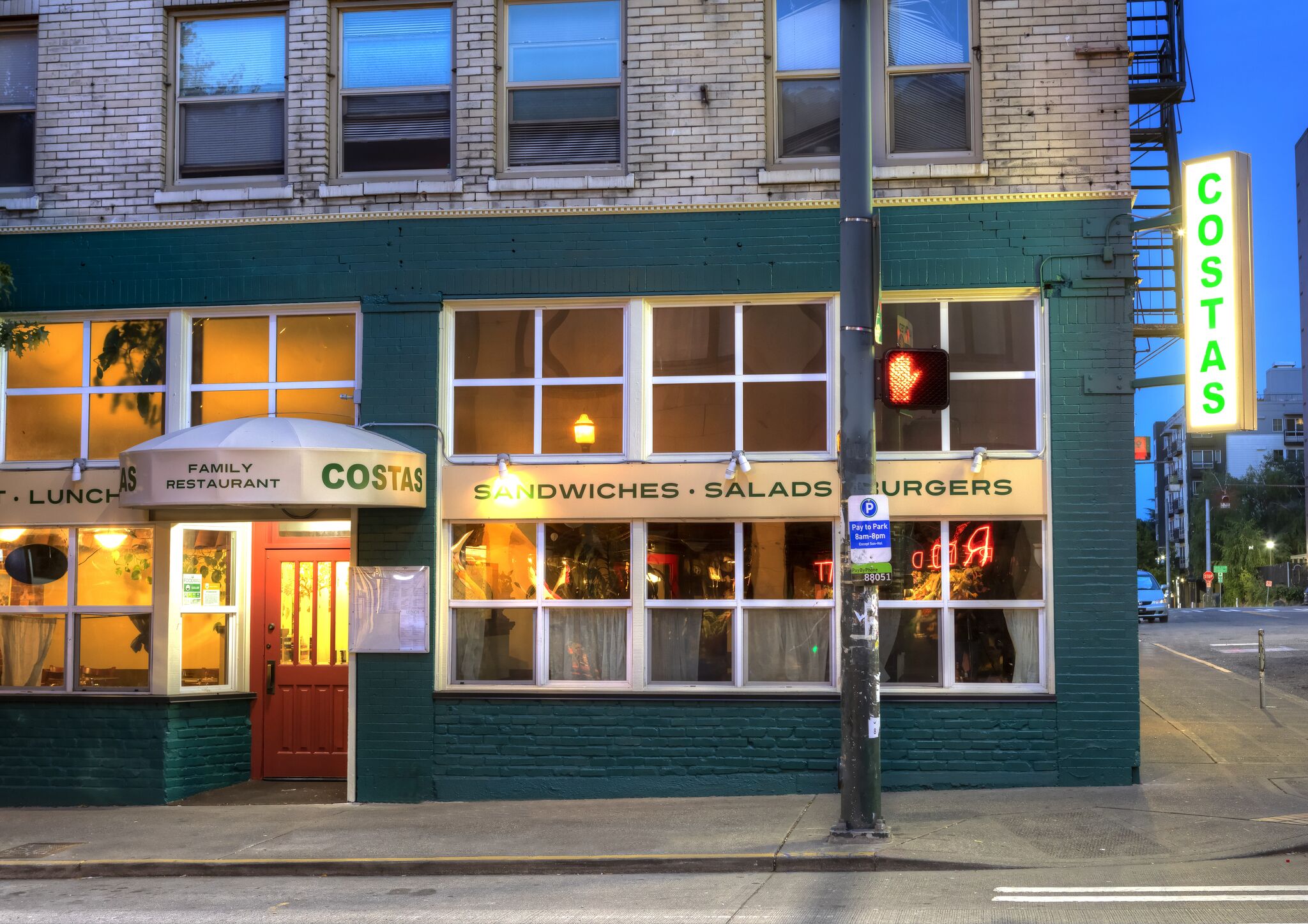 Street view of a two-story building with a sign reading "Costas" and windows displaying "Sandwiches, Salads, Burgers." A red stoplight is visible at the corner.