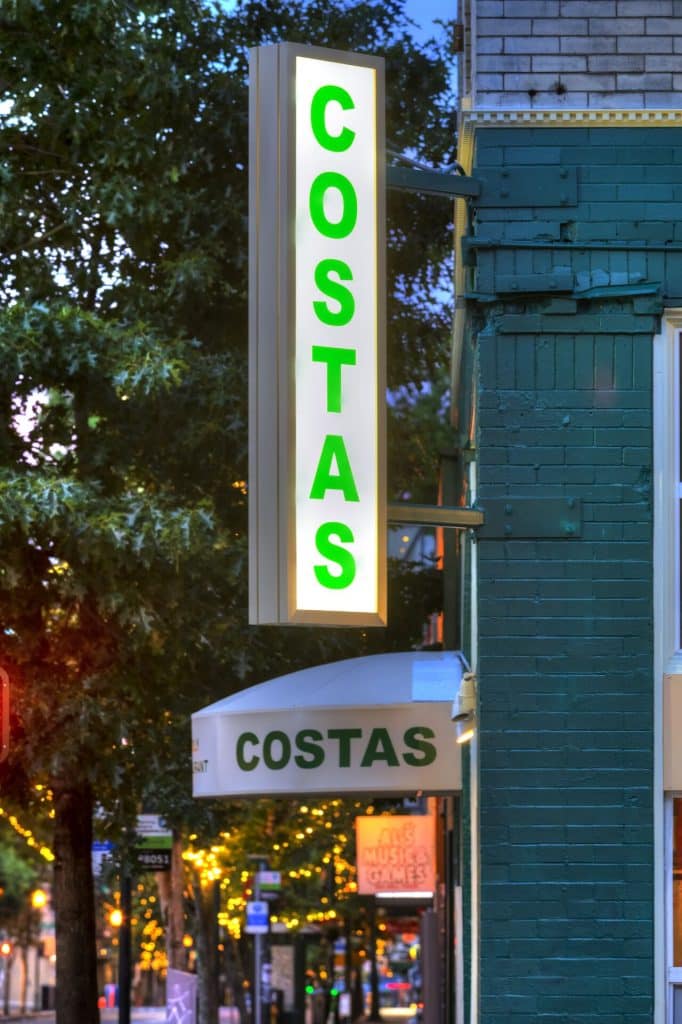 A vertical sign with green letters reading "COSTAS" on the side of a building, next to an awning with the same name. Trees and string lights are visible in the background.