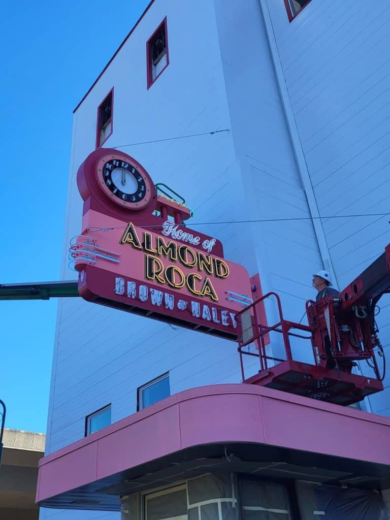 A worker in a lift installs a large pink sign saying "Home of Almond Roca, Brown & Haley" on a building, showcasing the sweet legacy celebrated by Almond Roca King across Seattle. A worker in a lift installs a large pink sign saying "Home of Almond Roca, Brown & Haley" on a building, showcasing the sweet legacy celebrated by Almond Roca King across Seattle.