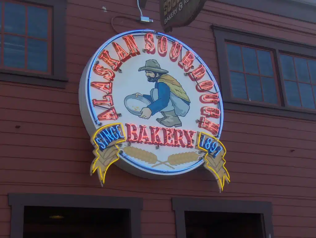 Sign for "Alaskan Sourdough Bakery" featuring a person kneading dough, with the text "Since 1899" on a building exterior. Sign for "Alaskan Sourdough Bakery" featuring a person kneading dough, with the text "Since 1899" on a building exterior.
