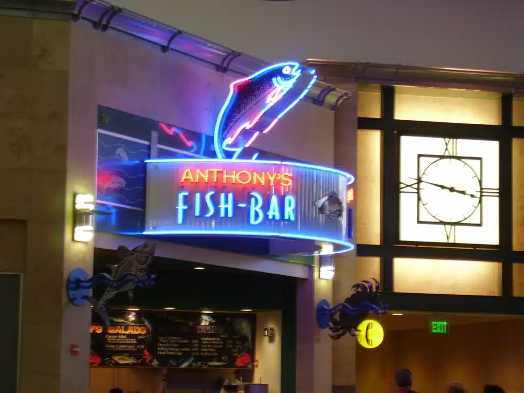 Neon sign reading "Anthony's Fish Bar" with a fish design, located indoors near a clock and illuminated exit sign. Neon sign reading "Anthony's Fish Bar" with a fish design, located indoors near a clock and illuminated exit sign.