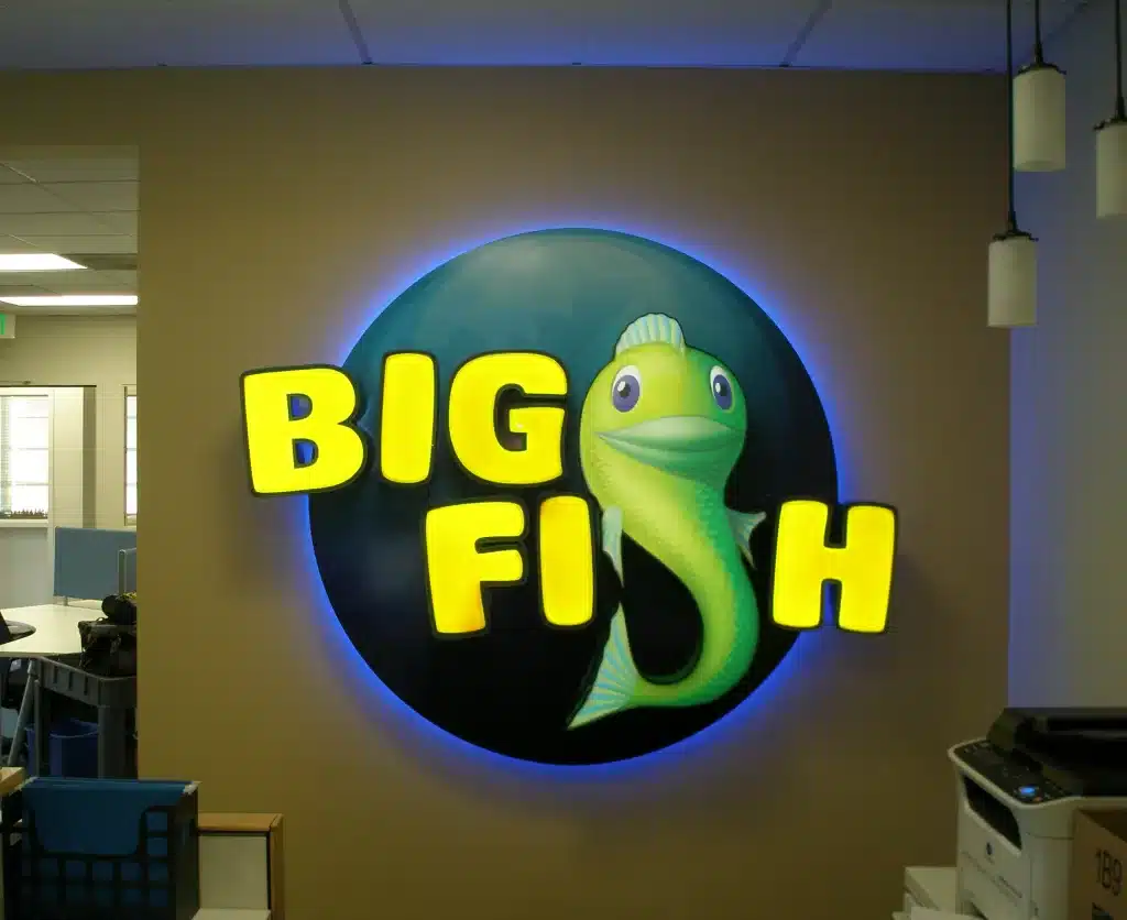 OLYMPUS DIGITAL CAMERA Illuminated "Big Fish" sign with a cartoon fish against a circular blue background on a beige wall.