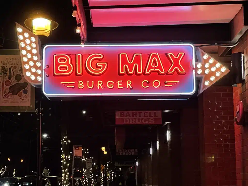 Neon sign reading "Big Max Burger Co" with illuminated arrows pointing towards the entrance, seen at night. Neon sign reading "Big Max Burger Co" with illuminated arrows pointing towards the entrance, seen at night.