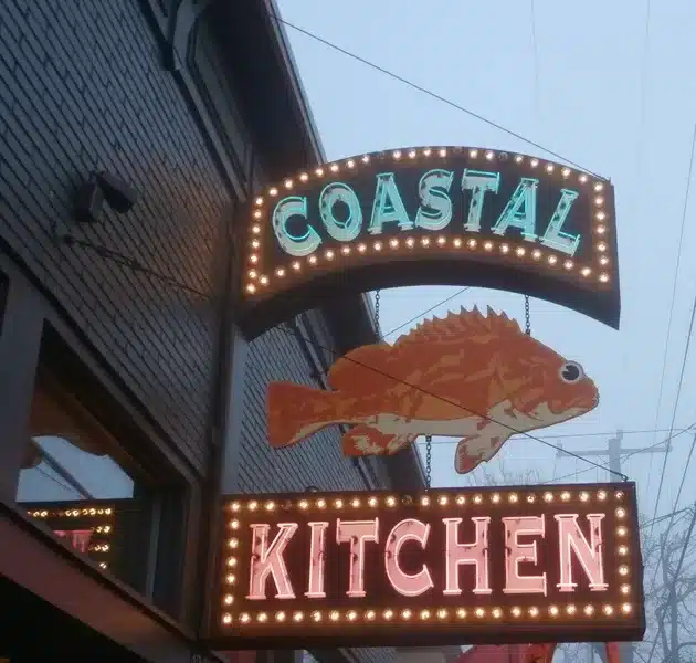 Sign for "Coastal Kitchen" with a lit-up fish graphic, attached to a brick building. Sign for "Coastal Kitchen" with a lit-up fish graphic, attached to a brick building.