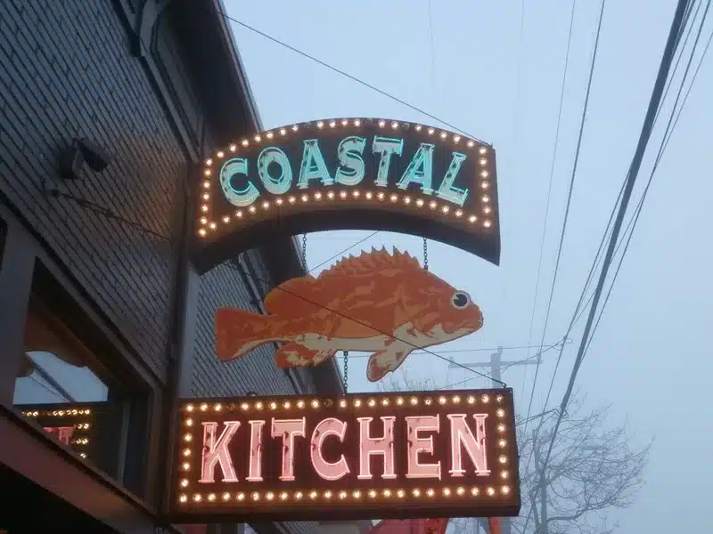 A lit sign displays a fish with "Coastal Kitchen" text below, attached to a building. A lit sign displays a fish with "Coastal Kitchen" text below, attached to a building.
