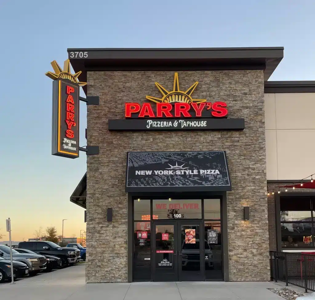 Exterior view of Parry's Pizzeria & Taphouse with a sign advertising New York-style pizza. The building is made of stone with large glass windows. Cars are parked nearby. Exterior view of Parry's Pizzeria & Taphouse with a sign advertising New York-style pizza. The building is made of stone with large glass windows. Cars are parked nearby.