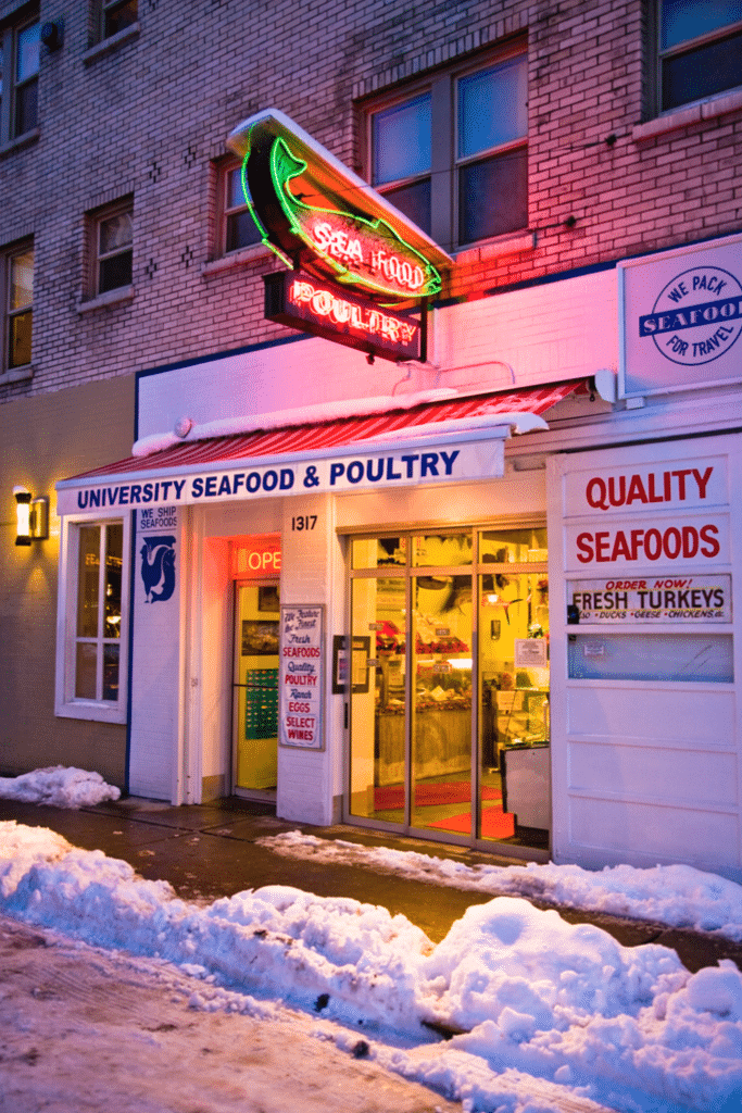 Street view of University Seafood & Poultry shop, showcasing Seattle's Neon signs gleaming against the backdrop of freshly fallen snow on the sidewalk.
