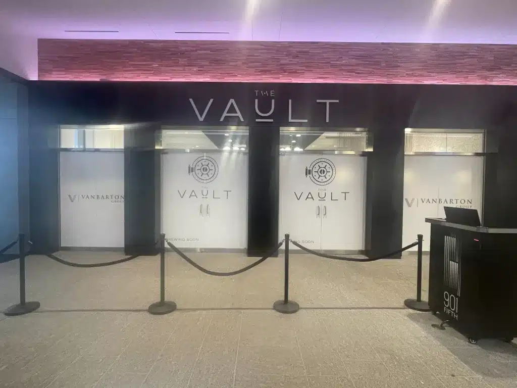 The Vault - Interior Sign A storefront with black and white signage reads "The Vault" and "Vanbarton." Three covered windows are labeled "Coming Soon." Velvet ropes are set in front.