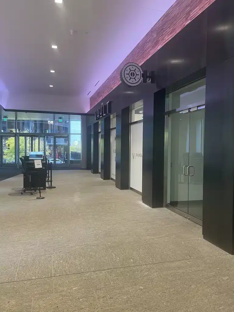 The Vault - Interior Sign 2 A modern, empty hallway with dark walls and a sign labeled "Vault." Glass doors and a reception desk are present, with natural light coming through large windows.