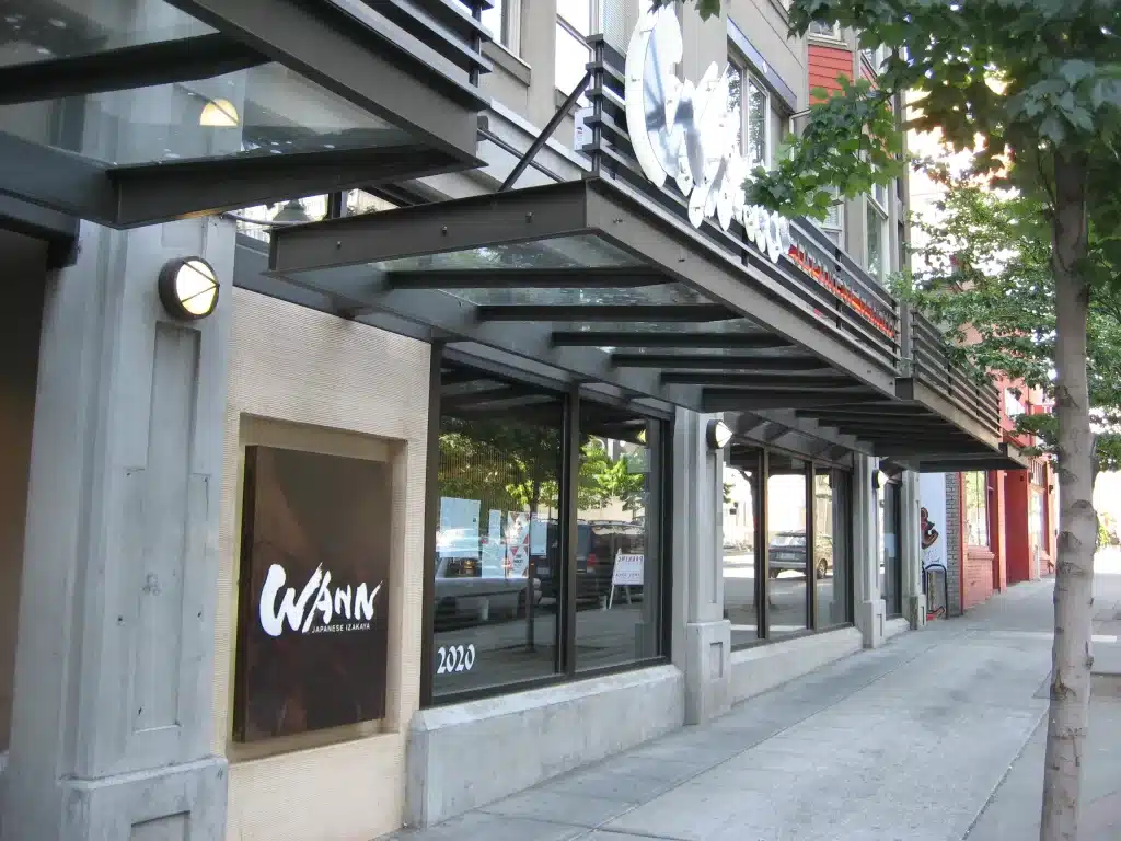 Street view of a modern building with large windows and a sign for "WANN Japanese Izakaya" on the facade. Trees line the sidewalk. Street view of a modern building with large windows and a sign for "WANN Japanese Izakaya" on the facade. Trees line the sidewalk.